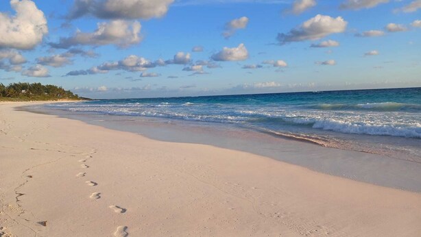A beach with footprints leading to the water, a clear sky, and a calm sea. - Eve's Cottage - Ocean Breeze Peaceful Getaway (Governor's Harbour)