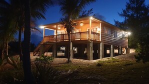 A two-story wooden house with a balcony, surrounded by palm trees and under a night sky. - Eve's Cottage - Ocean Breeze Peaceful Getaway (Governor's Harbour)