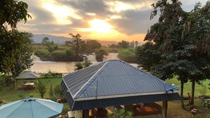 A rustic outdoor seating area with a table and chairs under a large umbrella, a small tent, and a view of a river at sunset. - Jangwani Camp Sagana (Murang'a)