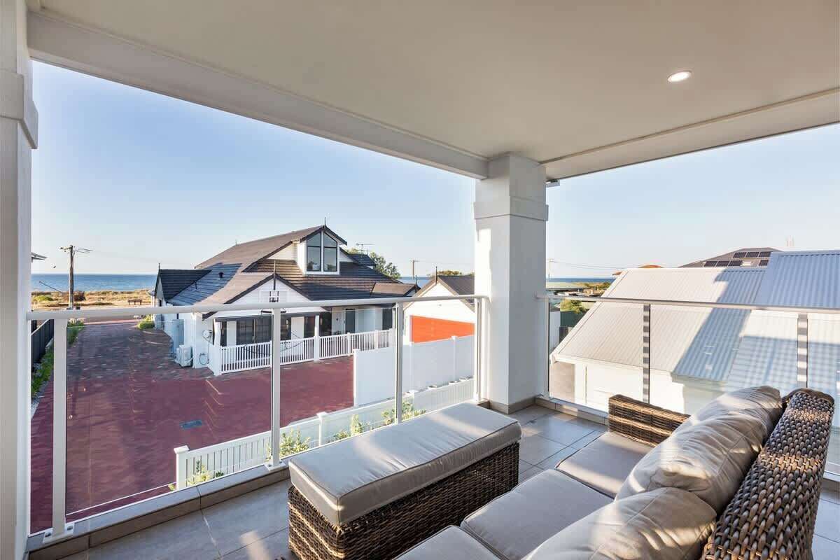 A balcony with a view of a beach and houses, wicker furniture, and a white railing.