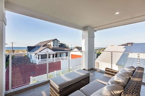 Un balcón con vistas a la playa y casas, mobiliario de mimbre y barandilla blanca.