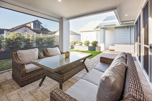 A covered patio with wicker furniture, a glass table, and a view of a house and garden.