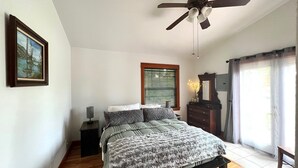 Ein Schlafzimmer mit Bett, Deckenventilator, Fenster mit Jalousien, Kommode und einem gerahmten Bild an der Wand.