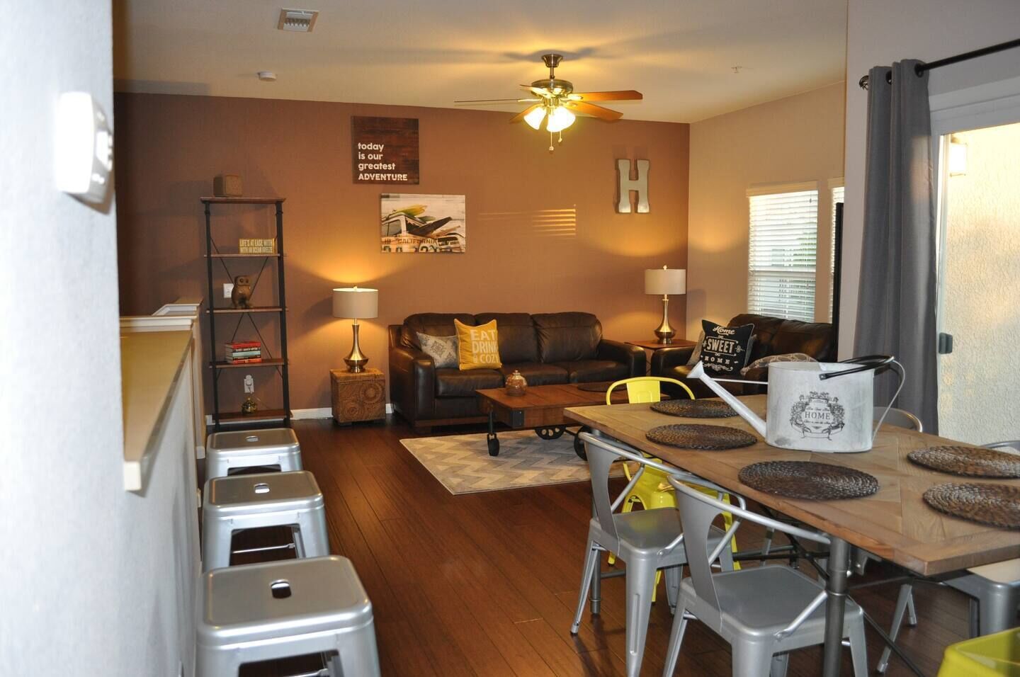 A living room with a sofa, coffee table, and a dining area with chairs and a table. There is a ceiling fan, a bookshelf, and a window with blinds.