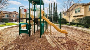 Un parque infantil con un tobogán y estructuras para escalar, rodeado de una zona de acolchado y edificios residenciales.