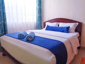 A neatly made bed with blue and white bedding, a blue pillow, and two rolled towels on a blue mat.