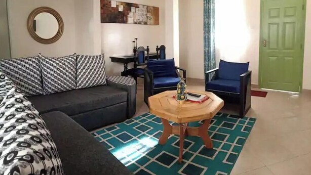 A living room with a sofa, a coffee table, and two armchairs. There is a green door and a window with curtains. - LUXE & SPACIOUS ONE BEDROOM UNIT (Kisumu)