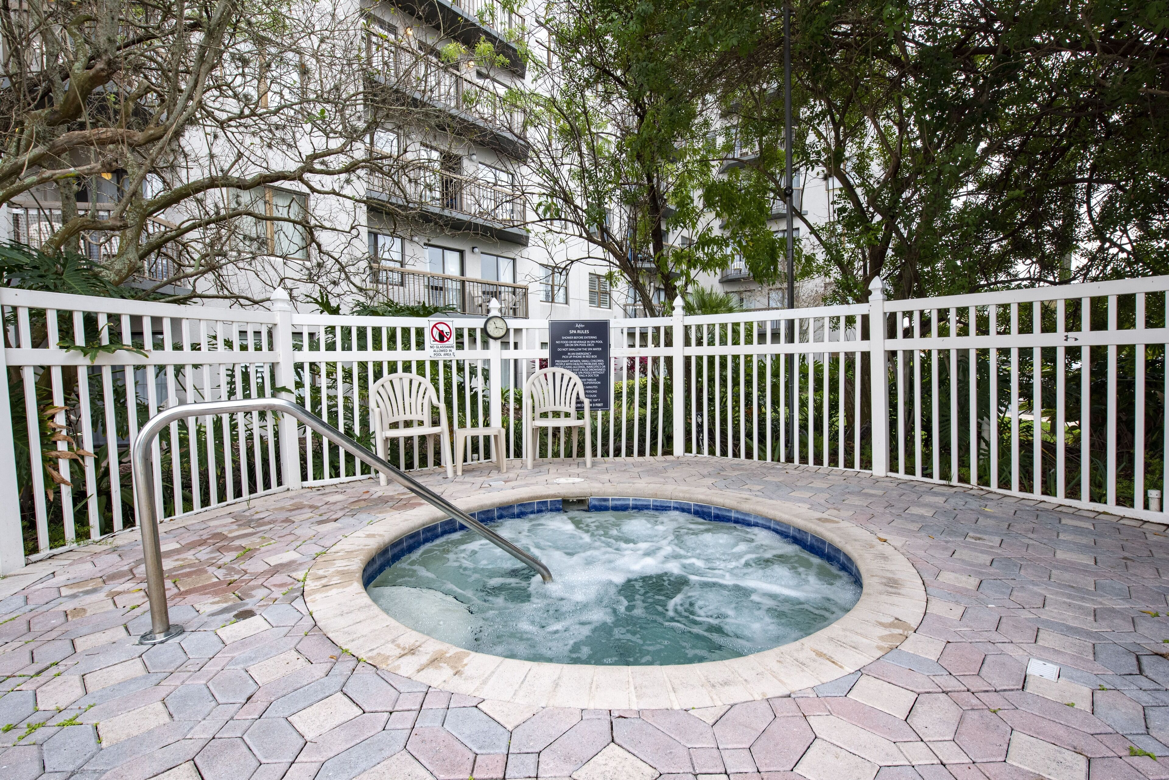 Un jacuzzi rodeado por una valla blanca, con un patio de ladrillo y árboles al fondo.