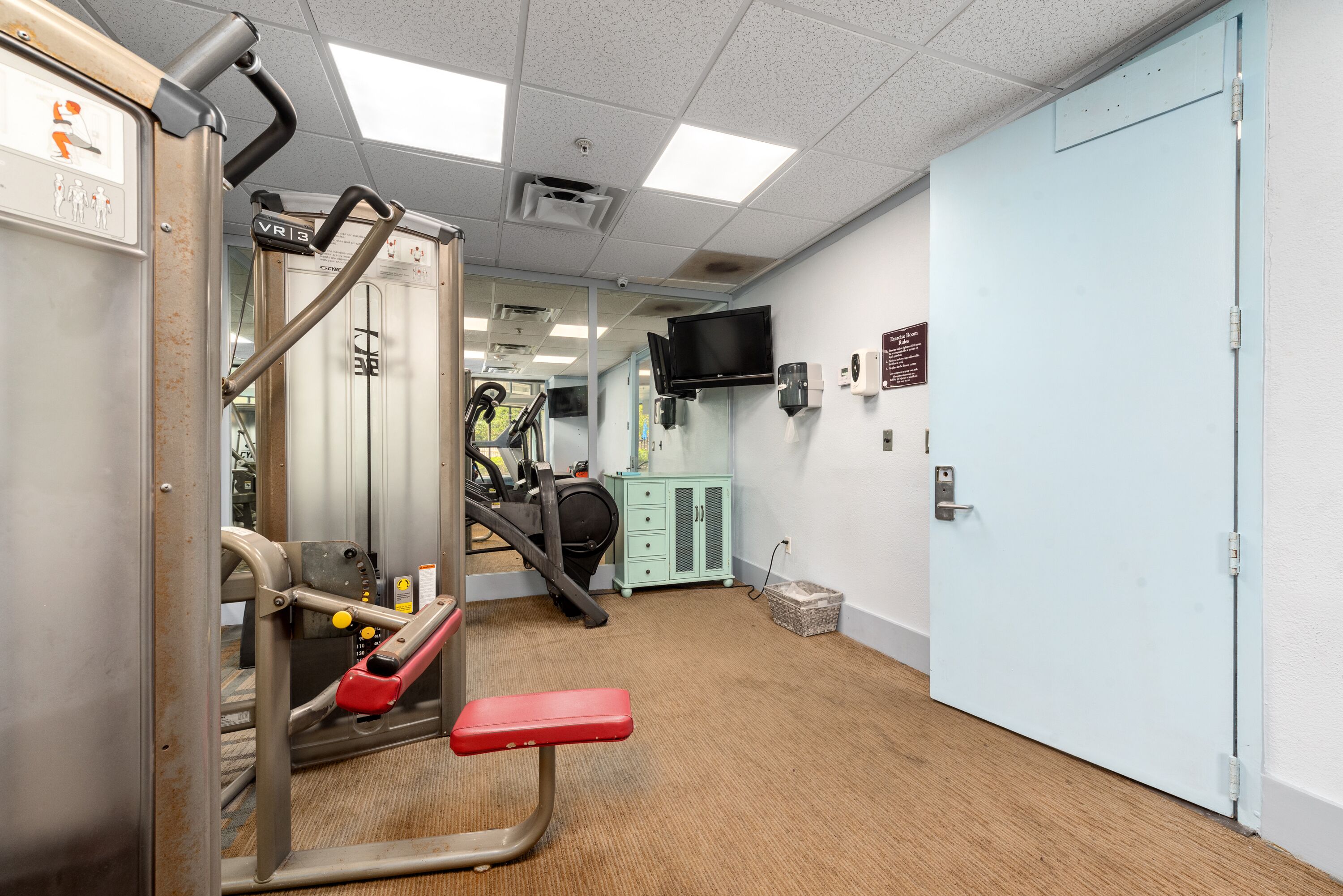 A gym with exercise equipment, a treadmill, a television on the wall, and a light blue door.