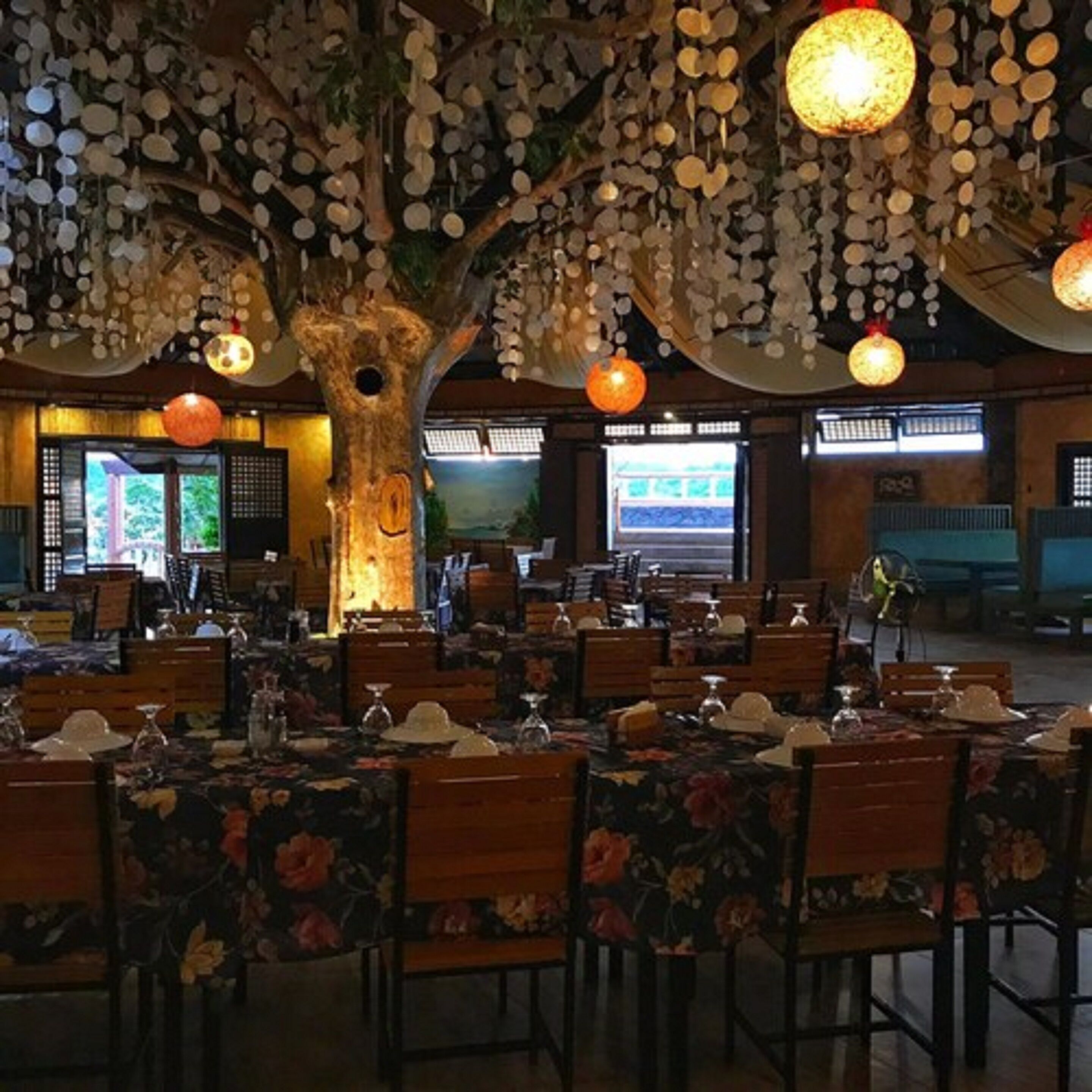 A restaurant with wooden tables and chairs, hanging light fixtures, and a large tree sculpture.