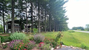 A residential area with a paved driveway, a flower bed with various plants, and a row of tall trees. - 4 bedrooms, 2 bathrooms and a three-season playroom,  Second-floor Unit. (Sturgeon Bay)