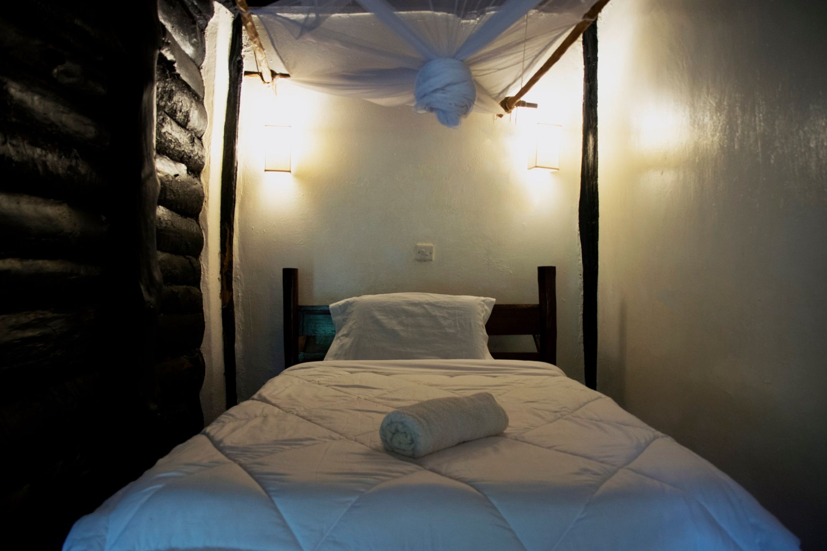 A single bed with a rolled towel, a wooden headboard, and two wall-mounted lights.