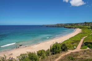 Une plage côtière offrant une vue dégagée sur l’océan, avec un rivage sablonneux et un sentier menant à un bâtiment.