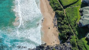 An aerial view of a beach with waves, a walking path, and a building with multiple roofs.