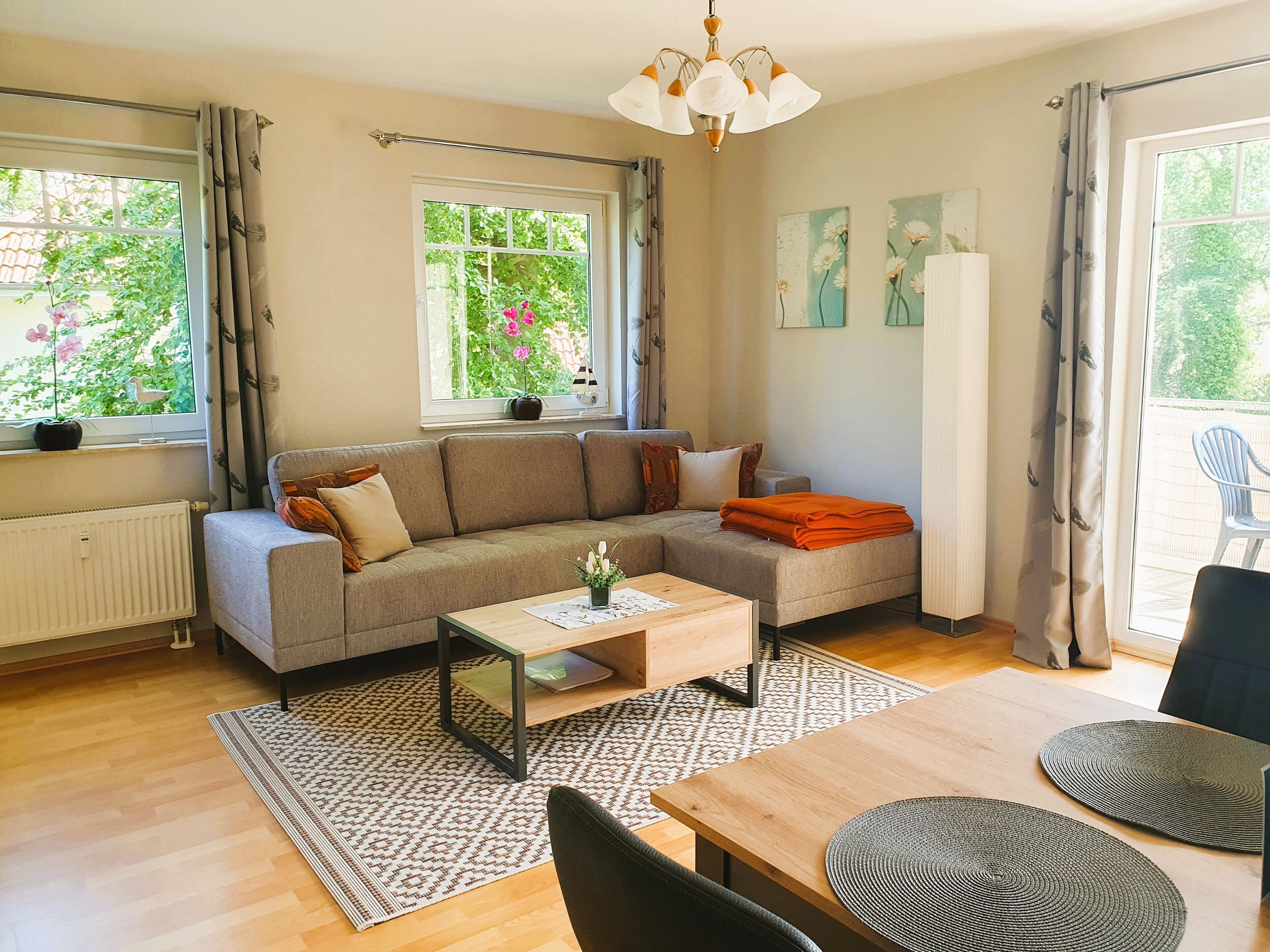 A modern living room with a sofa, coffee table, and dining table. There are windows with curtains, a radiator, and artwork on the walls.