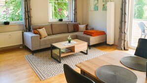 A modern living room with a sofa, coffee table, and dining table. There are windows with curtains, a radiator, and artwork on the walls. - Vacation apartment 7 Nachtigallenweg (Graal-Müritz)