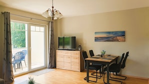 A room with a dining table, chairs, a TV on a wooden cabinet, a painting on the wall, and a view through a large window. - Vacation apartment 7 Nachtigallenweg (Graal-Müritz)