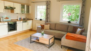 A modern kitchen with white cabinets, a built-in oven, and a coffee machine. There is a grey sofa with a patterned rug in front, and a window with curtains. - Vacation apartment 7 Nachtigallenweg (Graal-Müritz)