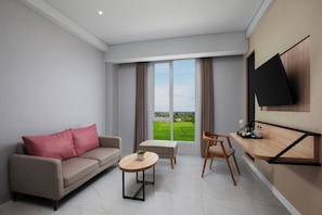 Un salon moderne comprenant un canapé, une petite table, un fauteuil, un téléviseur fixé au mur et une grande fenêtre donnant sur des champs verdoyants.