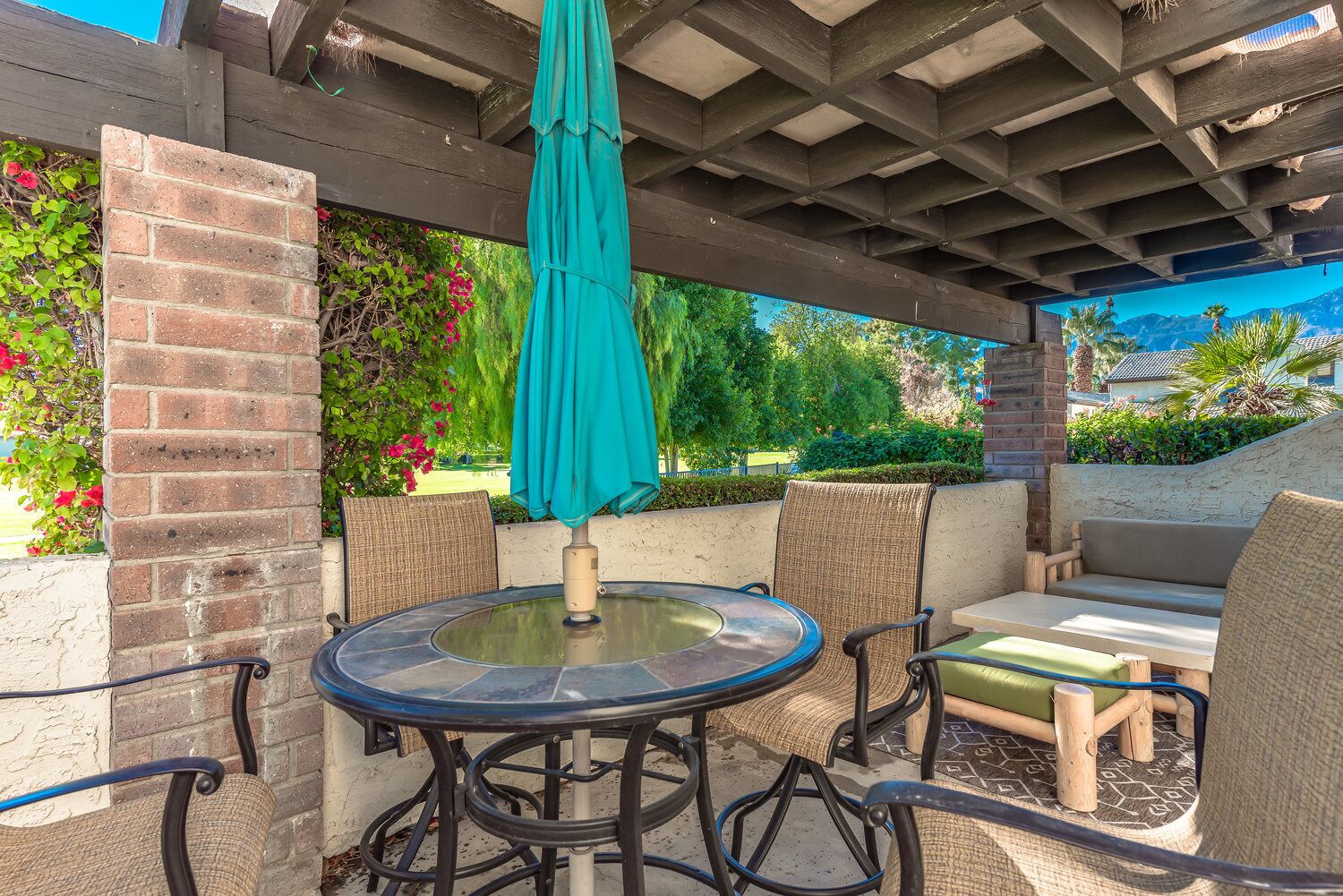 An outdoor patio with a round glass table, four wicker chairs, a teal umbrella, and a brick wall.