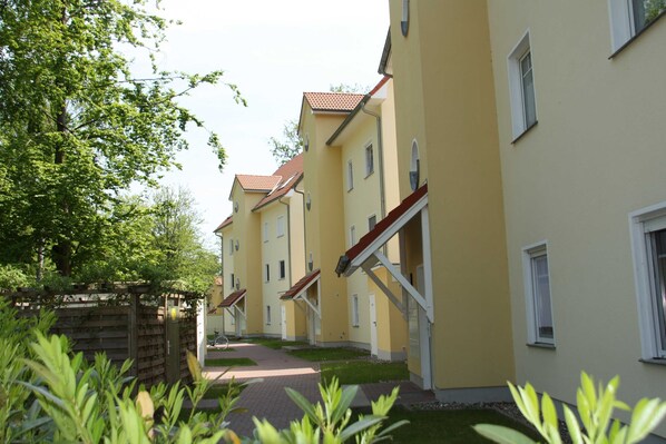 A row of yellow buildings with red roofs, a wooden fence, and green plants in the foreground.
