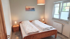 A bedroom with a wooden bed, two bedside tables with lamps, a painting on the wall, a window with blinds, and a radiator. - Vacation apartment Strandwiese 3 (Graal-Müritz)