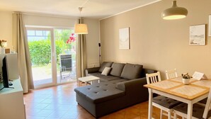 A modern living room with a grey sofa, a dining table, and chairs. There is a sliding glass door leading to a garden. - Vacation apartment Strandwiese 3 (Graal-Müritz)