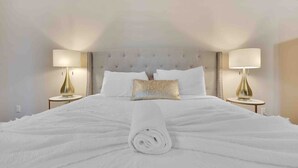 A neatly made bed with a rolled white towel in the center, flanked by two bedside tables with lamps, in a room with a tufted headboard and white walls. - Specials! Heated pool+spa|Game room|Sports courts (Thousand Palms)