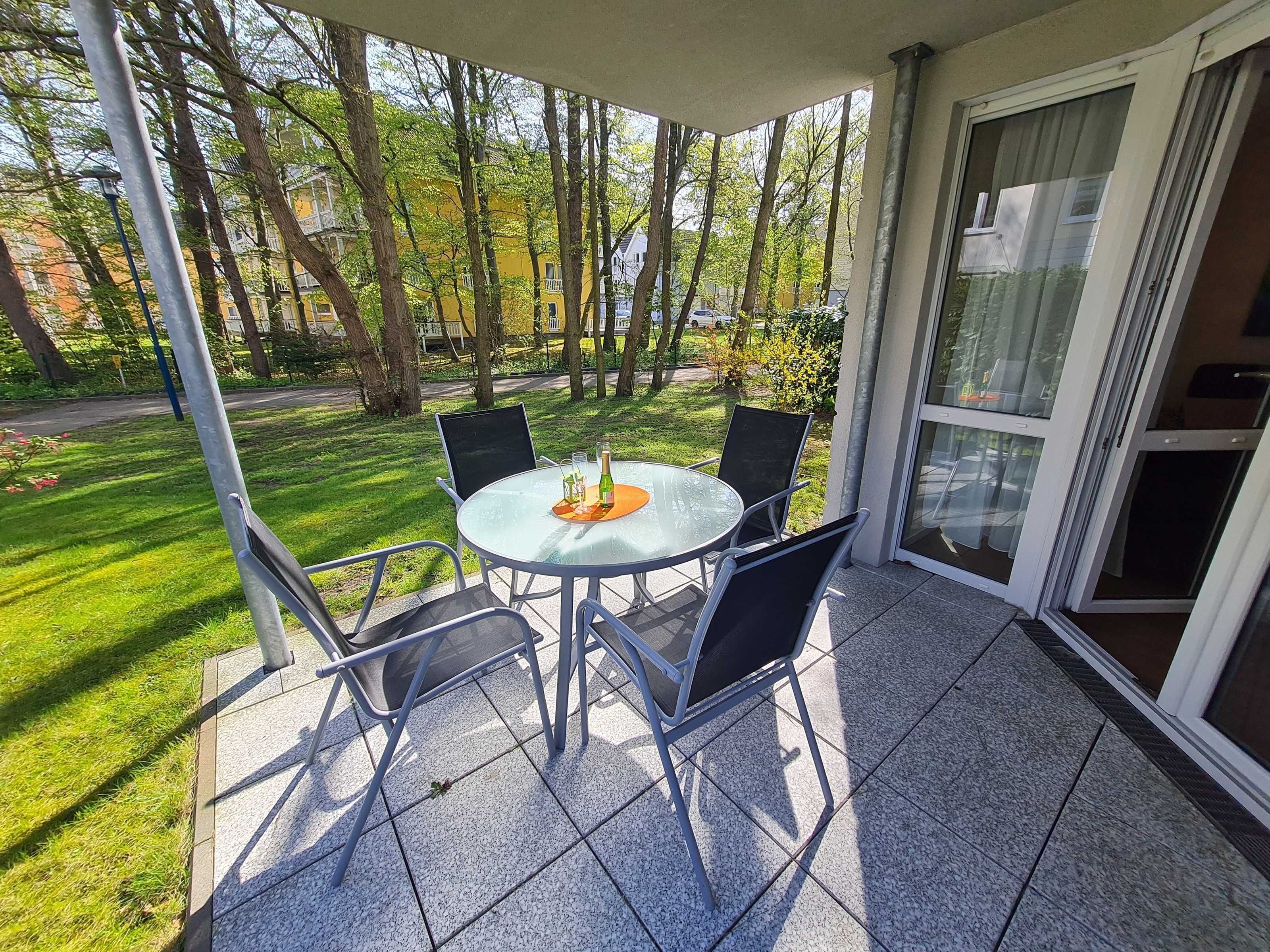 Een overdekt terras met een ronde tafel en vier stoelen, omgeven door een glazen deur en uitzicht op een groen gazon en bomen.