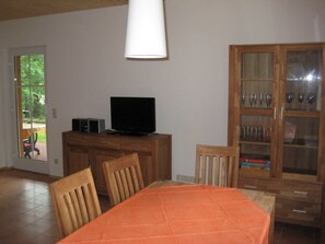 Un comedor con una mesa cubierta con mantel naranja, sillas de madera, un televisor sobre un mueble de madera y un aparador de vidrio con copas de vino.