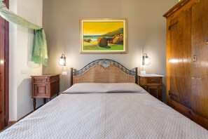 Un lit avec une tête de lit en osier, un tableau représentant un paysage côtier, une armoire en bois et une table de chevet avec une lampe.
