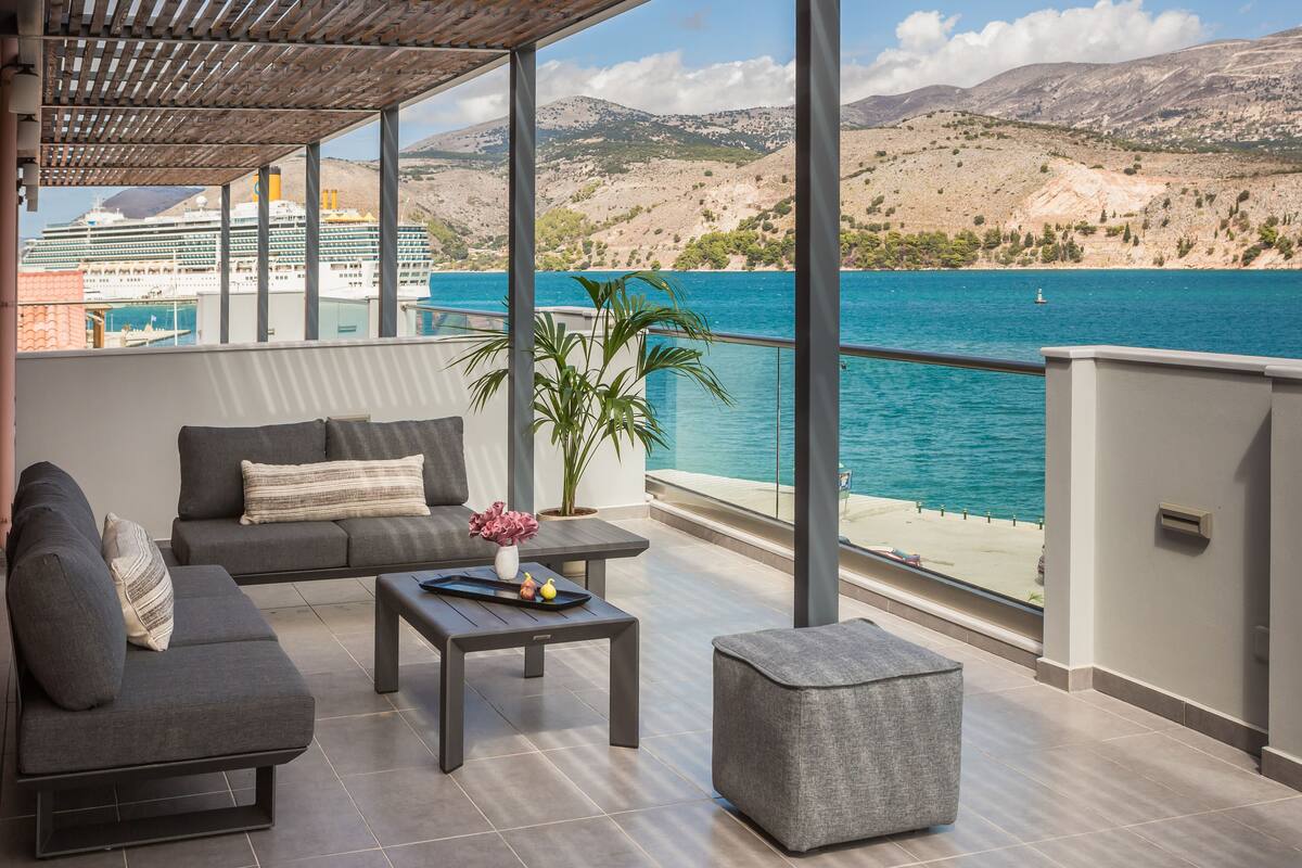 A modern balcony with a sofa, a small table, and a potted plant, overlooking a body of water and a cruise ship.