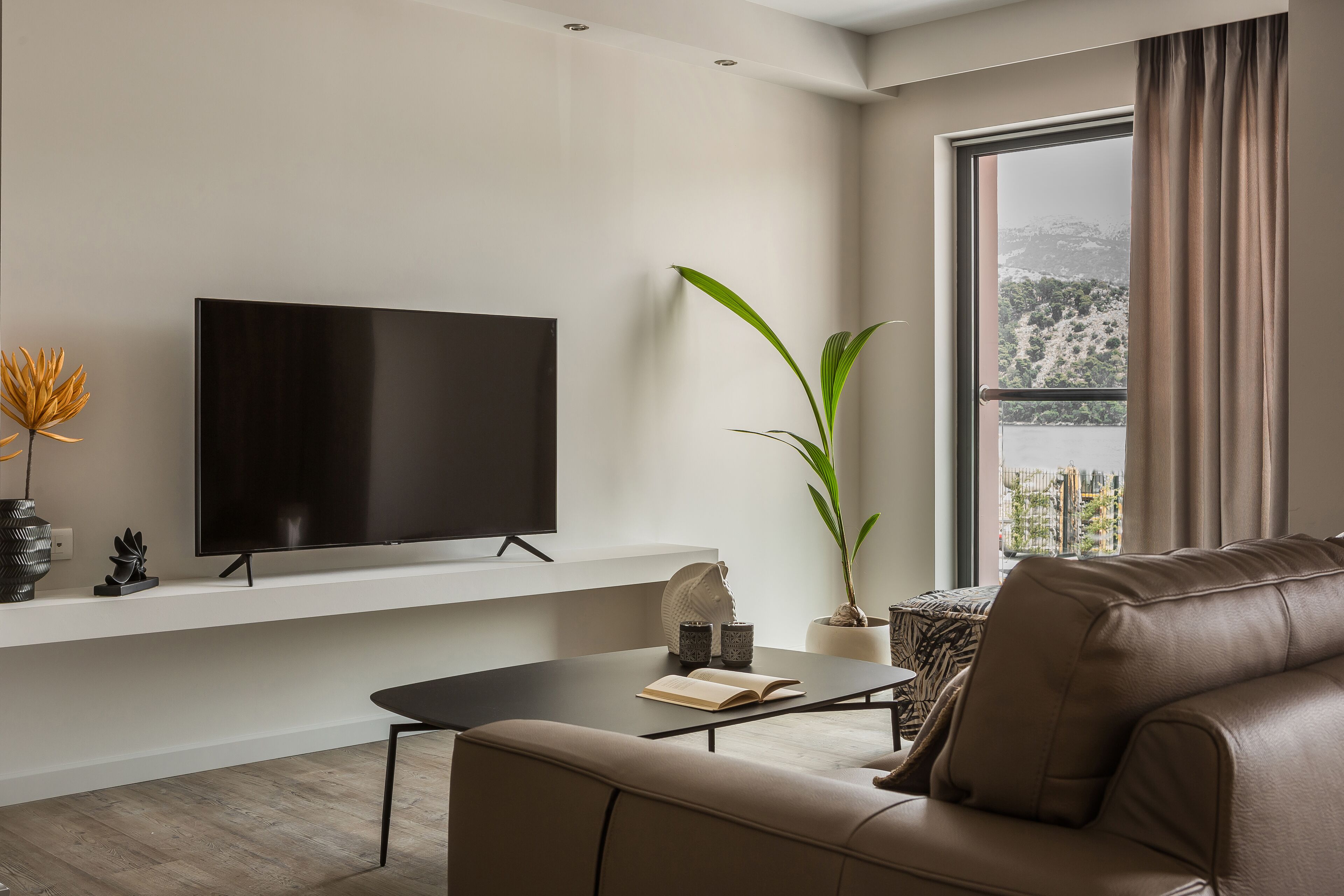 Un salon moderne doté d’un téléviseur à écran plat, d’un canapé en cuir marron, d’une table basse et d’une grande fenêtre offrant une vue sur un paysage montagneux.