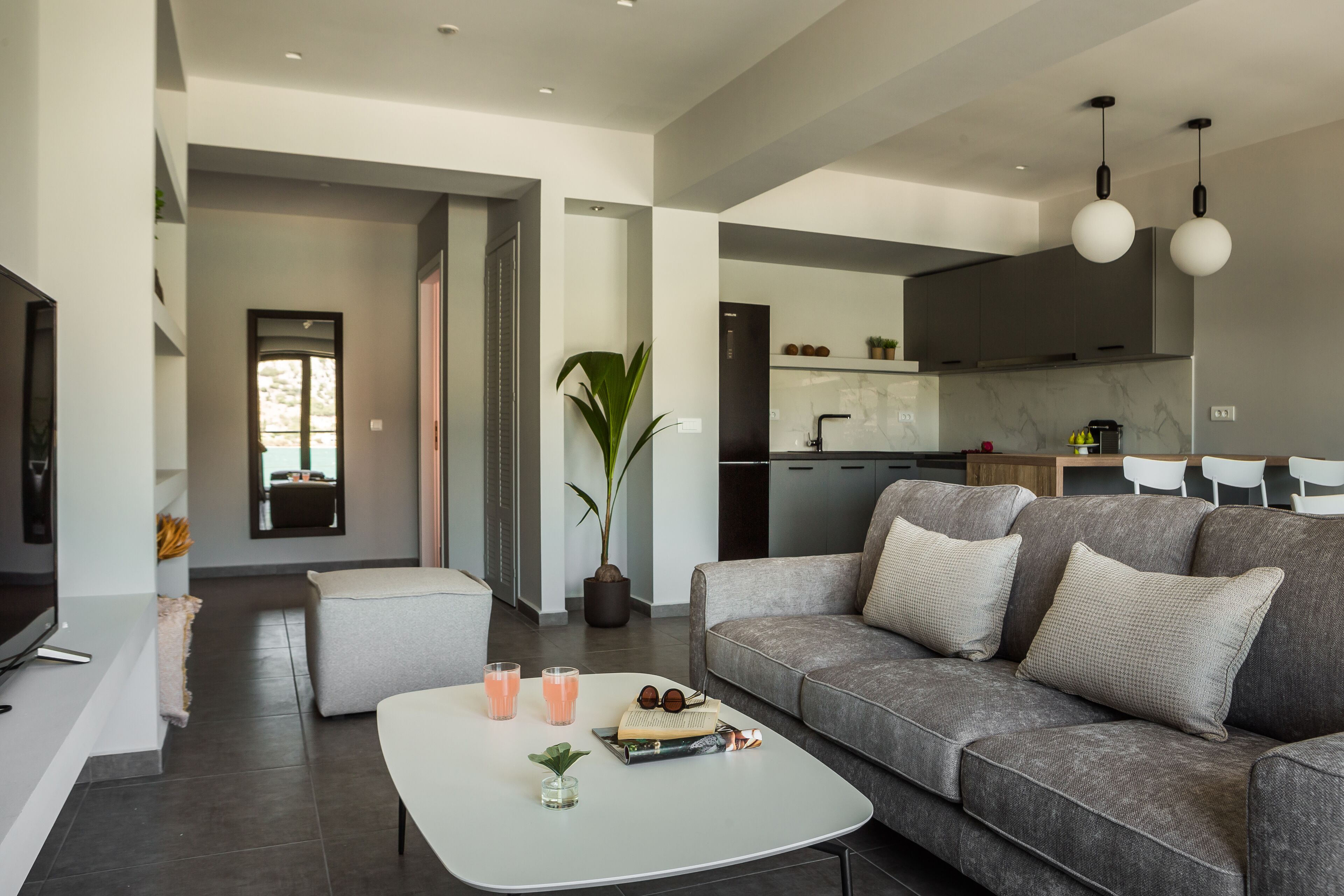 A modern living room with a grey sofa, a white coffee table, and a TV. There is a kitchen in the background with white cabinets and a marble backsplash.
