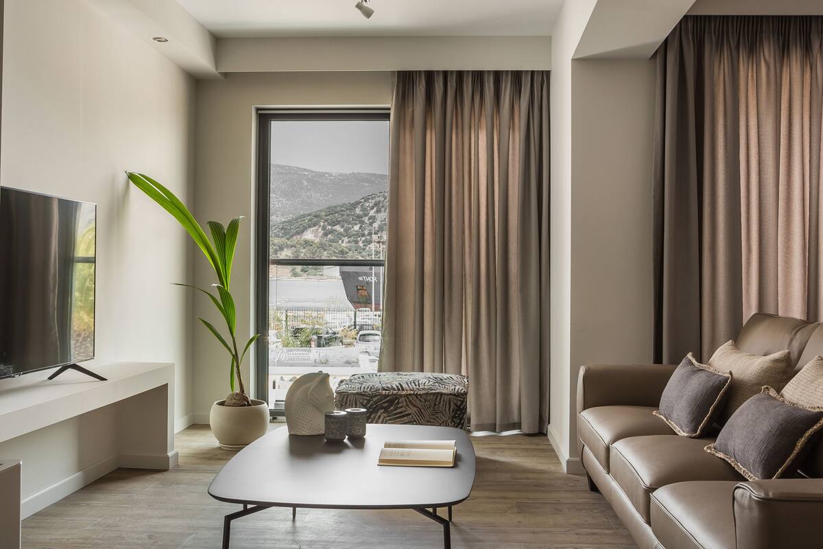 A modern living room with a flat-screen TV, a potted plant, a sofa, and a view of a mountainous landscape through a large window.