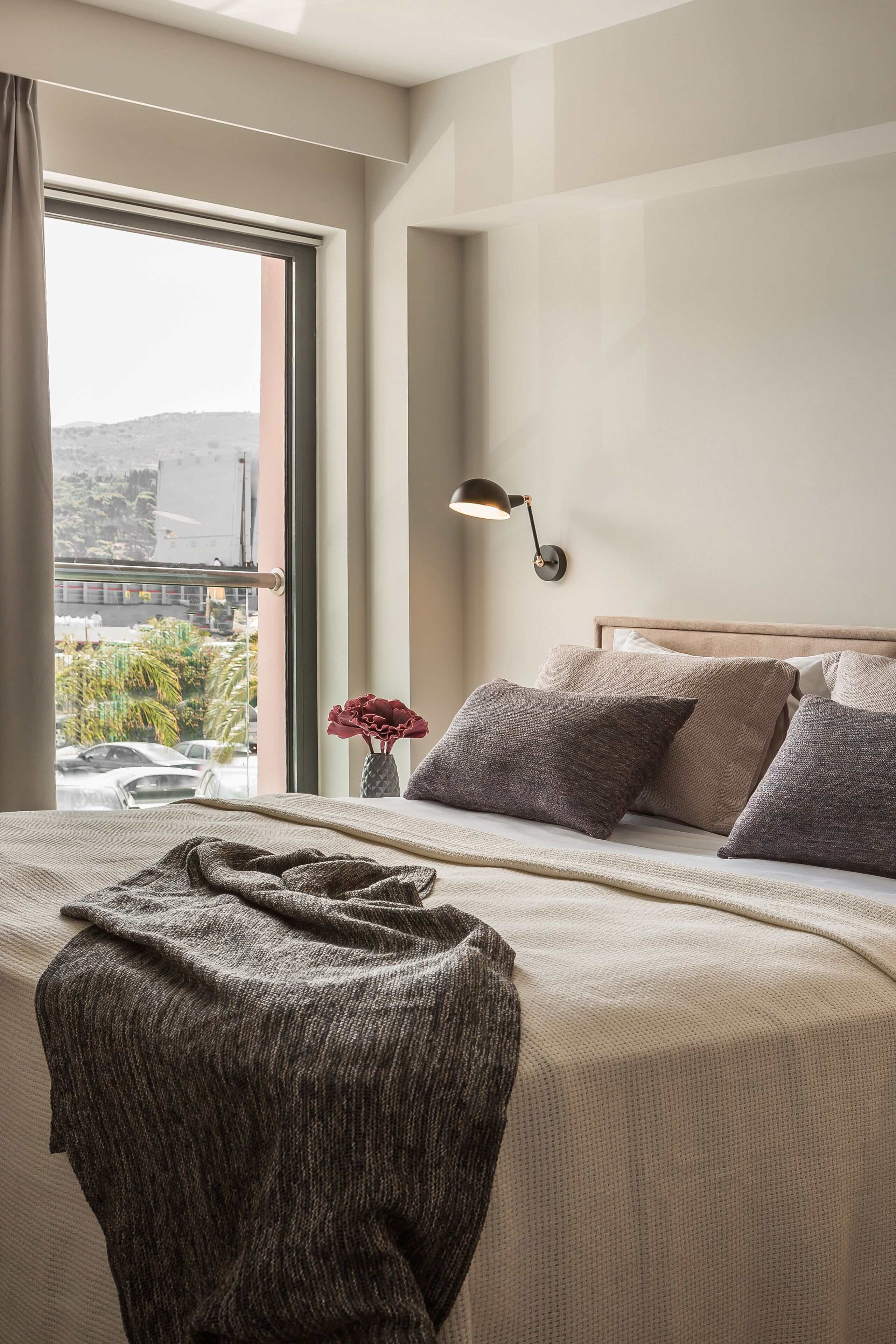 A hotel room with a large bed, a bedside lamp, and a view of a cityscape through a large window.