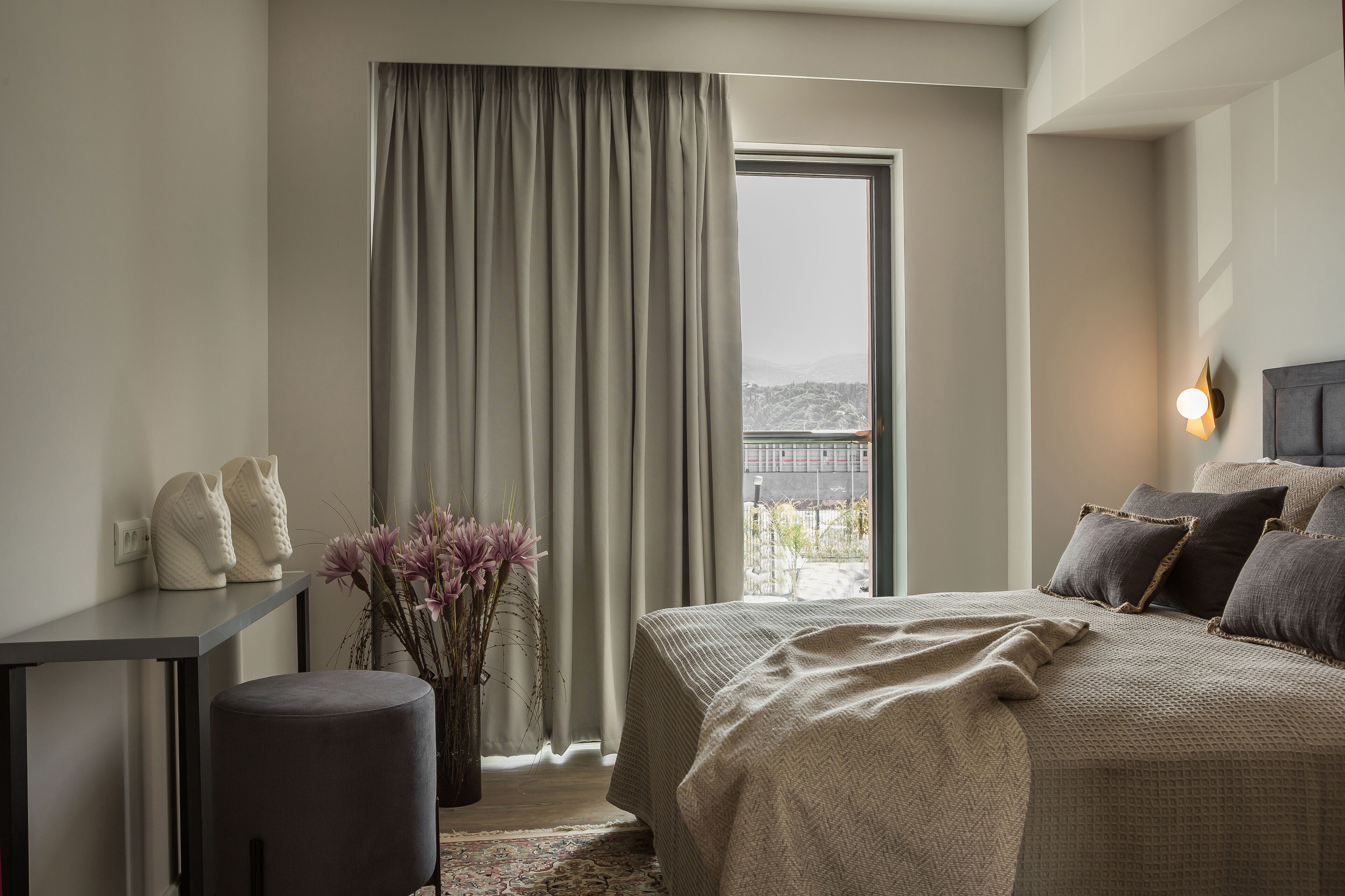 Ein Schlafzimmer mit einem Bett, einem Schreibtisch, einem Sessel, einer Vase mit Blumen und Blick ins Freie durch ein Fenster mit Vorhängen.