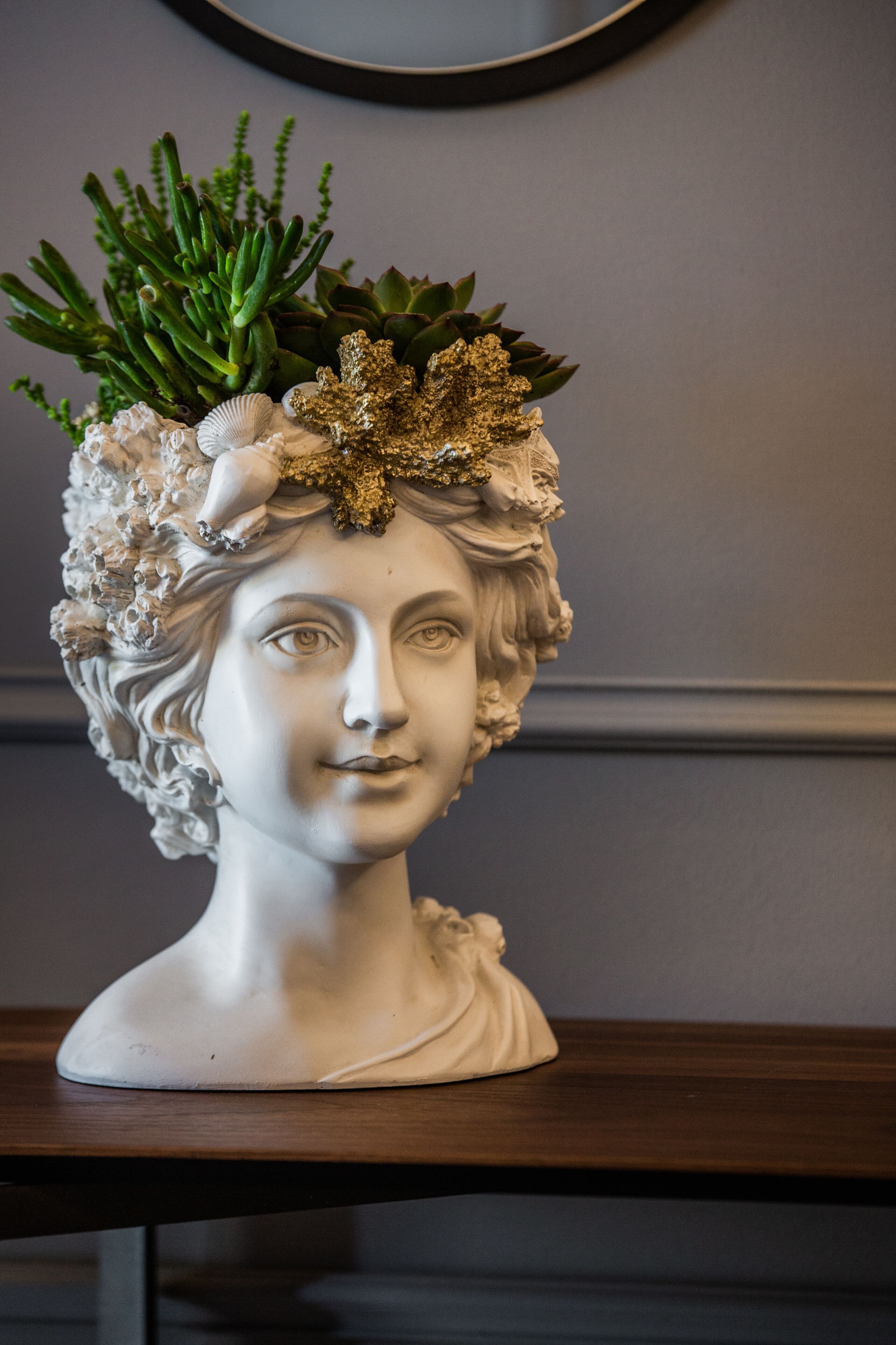 A bust sculpture with a floral arrangement on top, placed on a wooden surface against a plain wall.