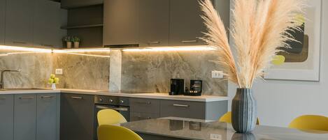 A modern kitchen with grey cabinets, a marble countertop, and a dining area with yellow chairs.