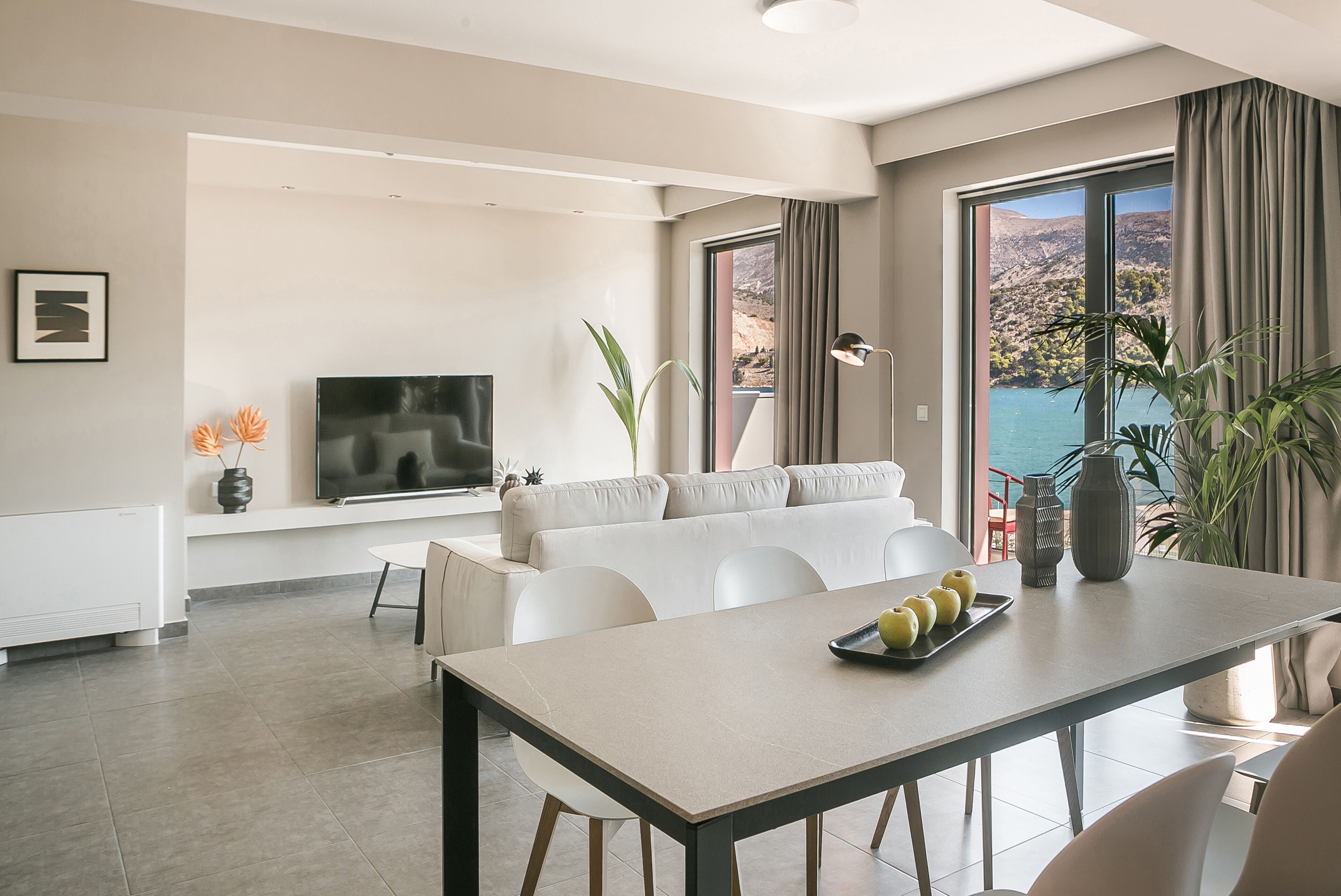 A modern living room with a sofa, dining table, and a TV. There are plants and a view of a mountainous landscape through large windows.