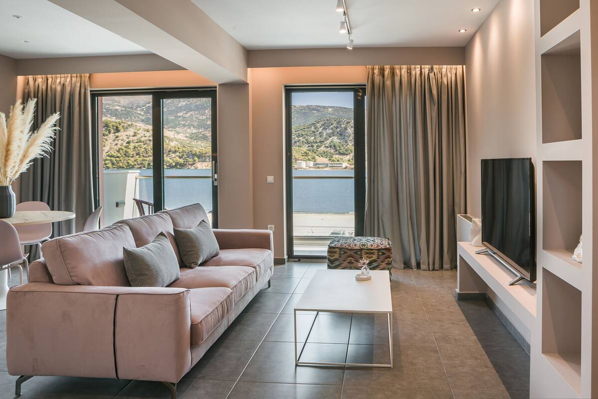 A modern living room with a sofa, coffee table, and TV. Large windows offer a view of the outdoors.