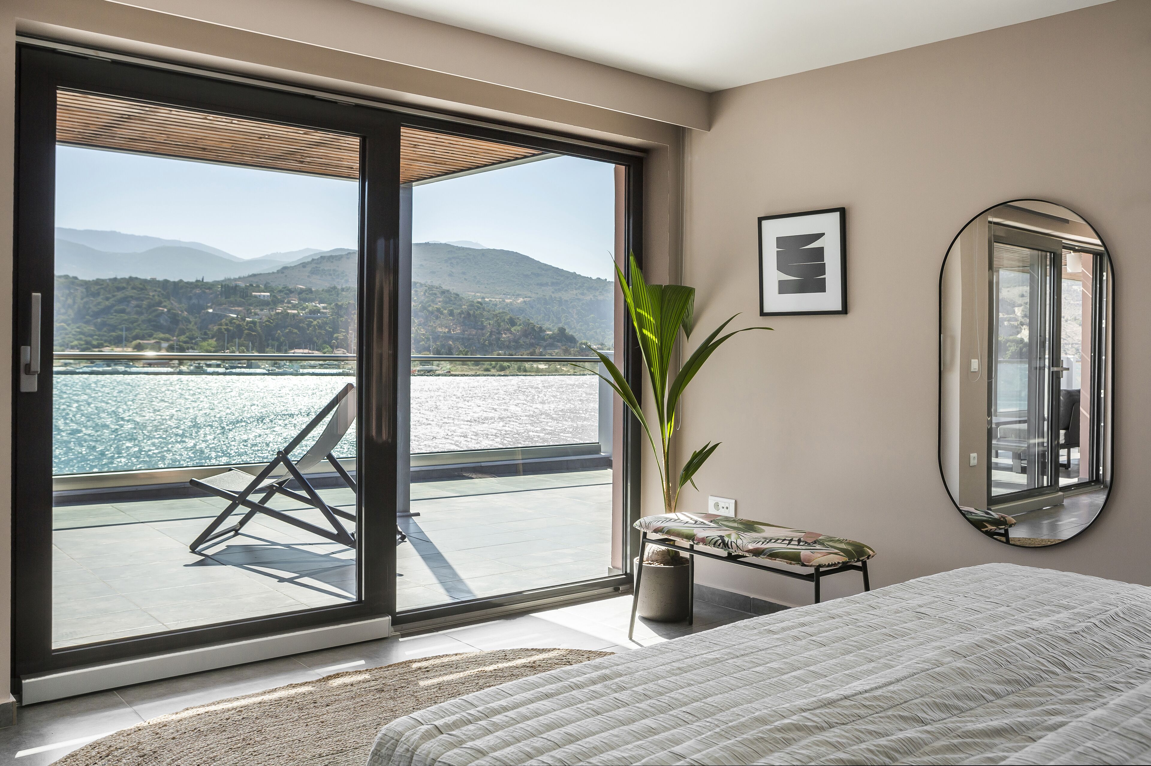 A bedroom with a large sliding glass door leading to a balcony with a view of a body of water and hills.
