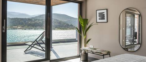 A bedroom with a large sliding glass door leading to a balcony with a view of a body of water and hills.