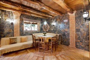 A stone-walled room with a wooden ceiling, a stone bench, a round table with chairs, and a window with a view.