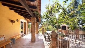 A covered patio with brick flooring, wooden benches, a brick barbecue, and a metal fence.