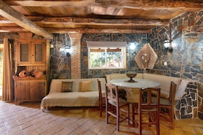 A rustic room with a stone wall, wooden ceiling beams, a stone fireplace, a wooden cabinet, a sofa, and a round table with chairs. - 2-bed Holiday Apartment with Pool, Wi-Fi, Garden & Terrace (Armação de Pêra)