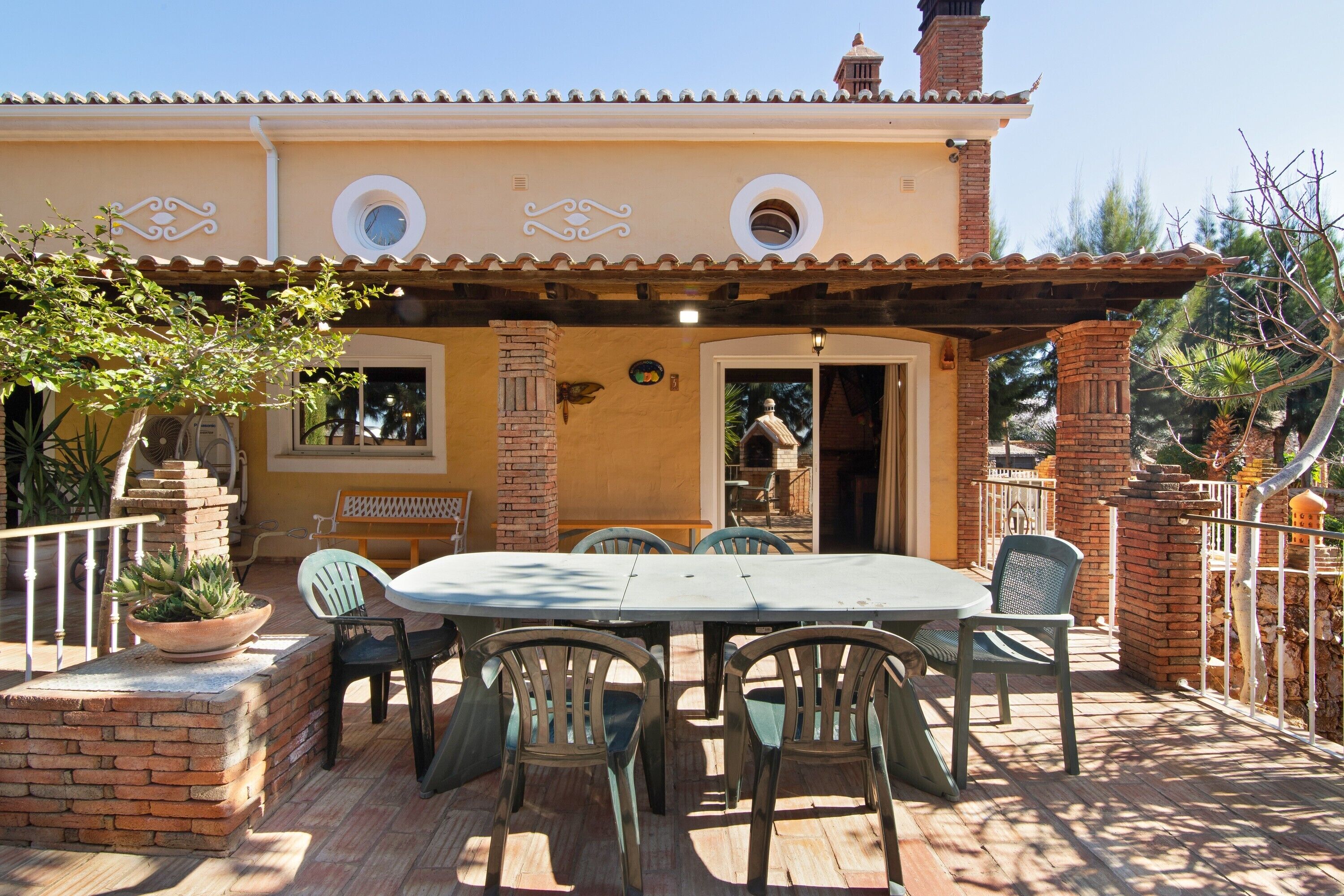 Un patio con una mesa y sillas, una jardinera de ladrillo con plantas y un edificio con tejado de azulejos y detalles decorativos.