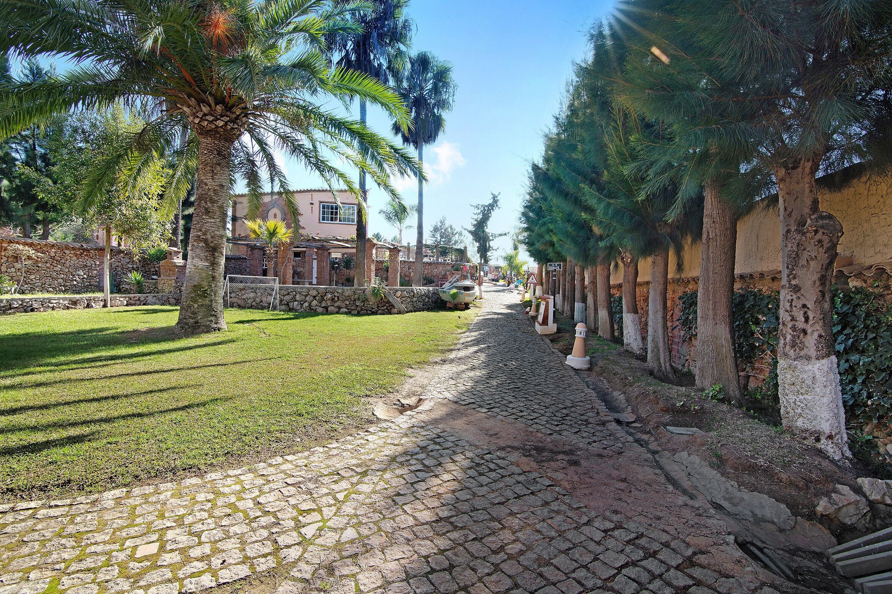 A cobblestone pathway flanked by trees and a stone wall, leading to a building with a stone facade.