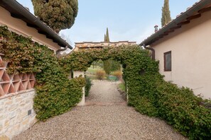 A pathway leading through an archway covered in greenery, flanked by buildings with stone walls and tiled roofs.