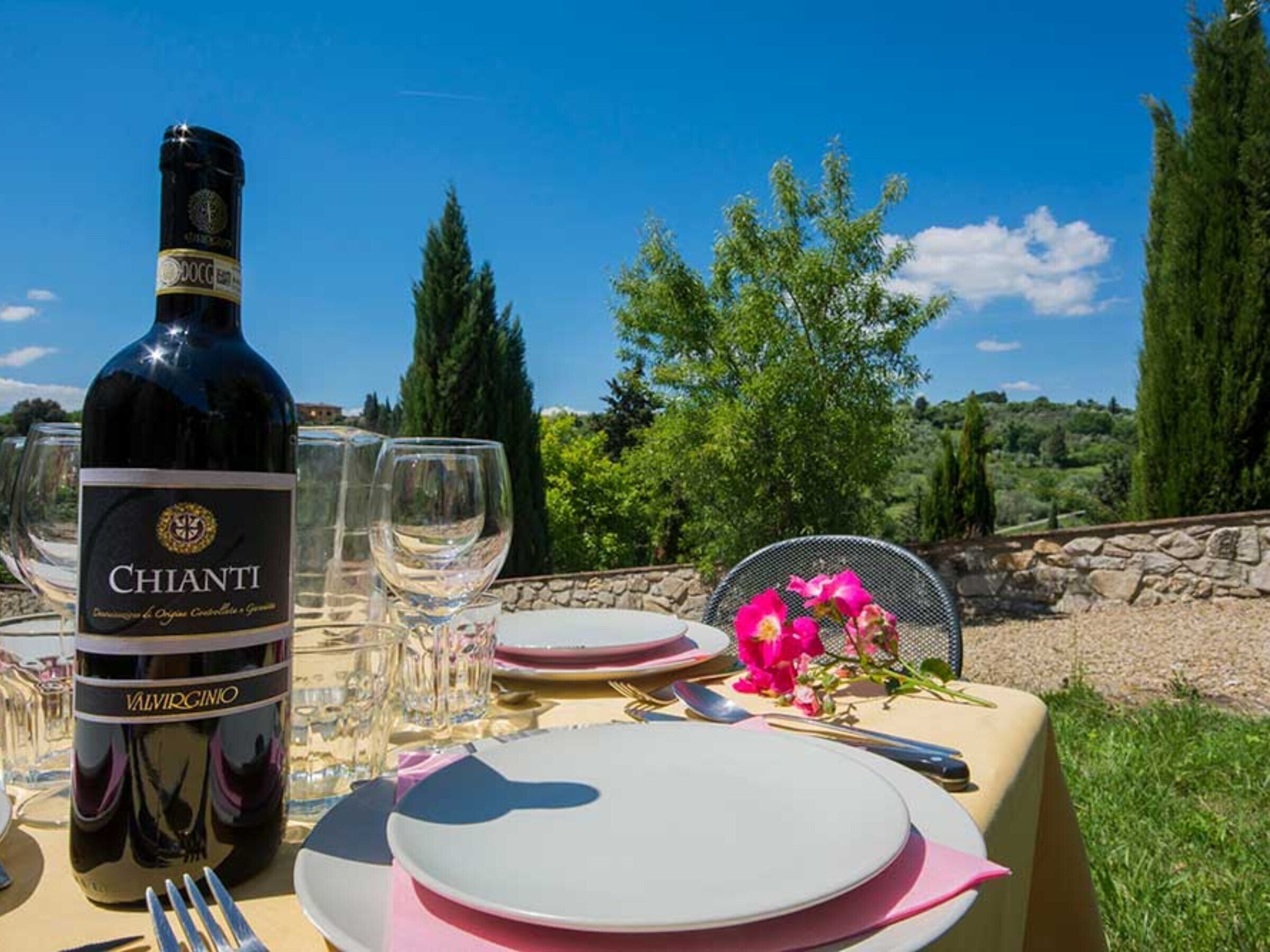 Une table dressée à l’extérieur, avec une bouteille de vin Chianti, des verres, des assiettes et une chaise agrémentée d’une fleur.